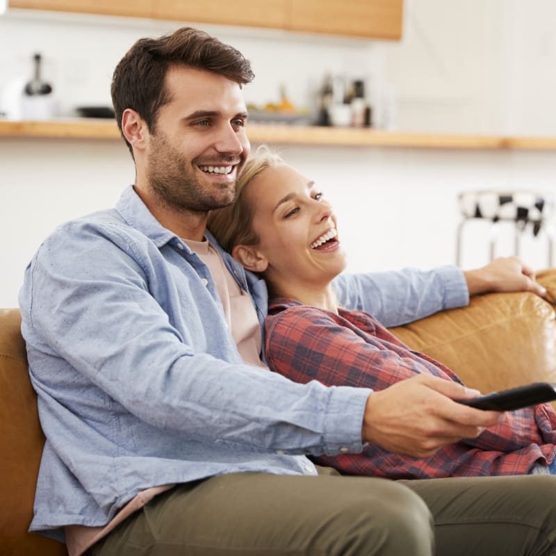 Man and woman smiling together, with the woman hugging him from behind while he holds her hands.