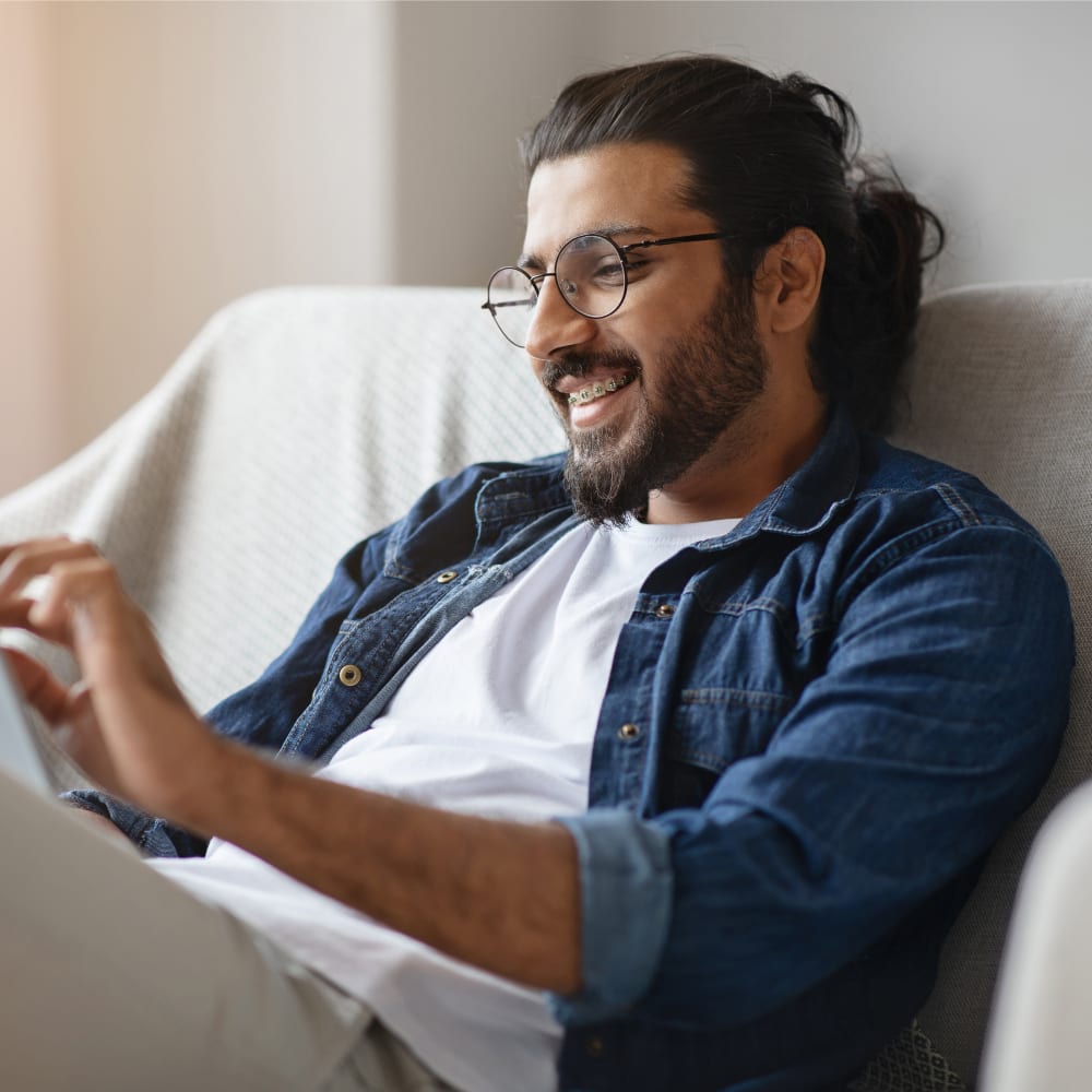 A man is sitting on a couch using a tablet. He has round glasses, and his smile shows braces.