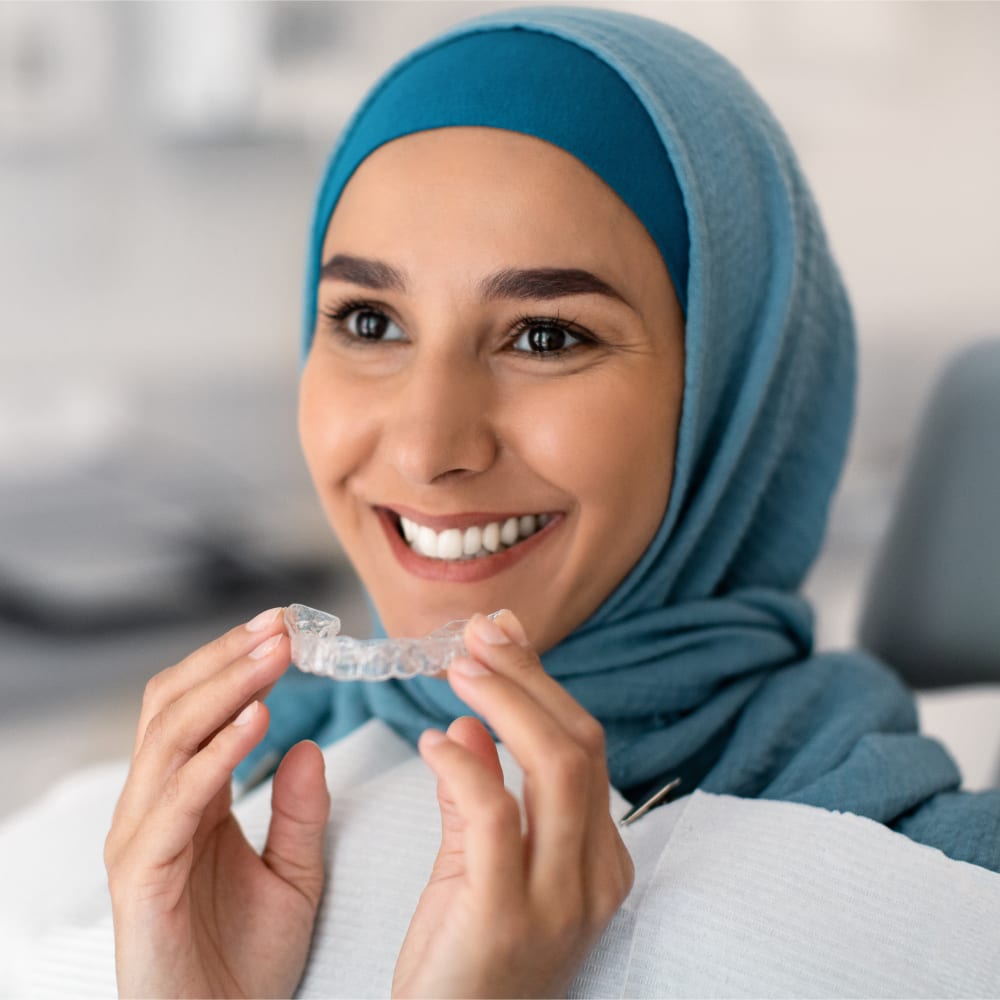 A woman in a blue head scarf is smiling and holding a clear aligner tray.