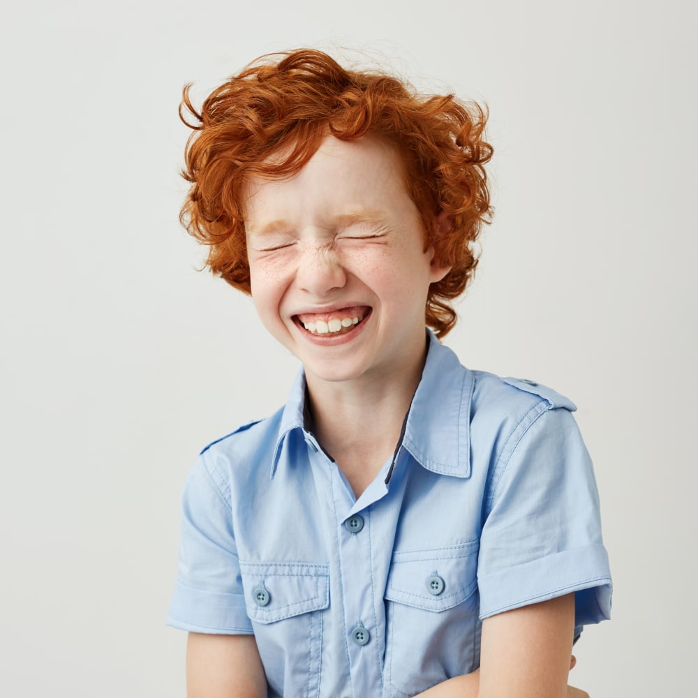Boy with red hair wearing a blue shirt smiling at the camera with his eyes slightly squinted.