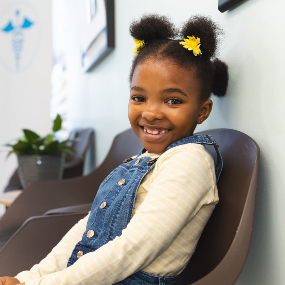 Young girl with curly hair wearing a yellow top and blue denim overalls, smiling at the camera.