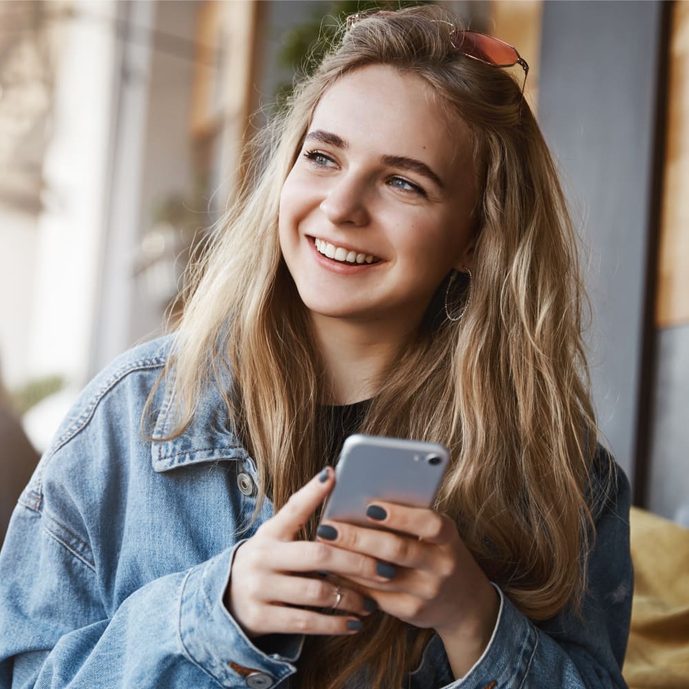 Young woman with blonde hair wearing a blue denim jacket, smiling while holding a cellphone.