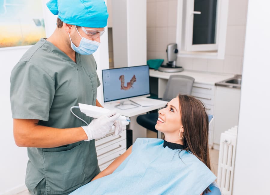 A dentist and a patient discuss oral health. A digital rendering of the patient's teeth is on the screen behind them.
